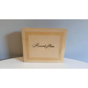 May include: A vintage record album with a cream-colored cover. The cover features a decorative border and the words "Record Album" in elegant script. The album is rectangular and appears to be made of a textured material, suggesting a classic design.