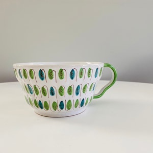 以下が含まれることがあります： 緑色の取っ手と緑と青の葉の模様がついた白いセラミック製のマグカップです。