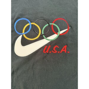 Może przedstawiać: Czarny t-shirt z logo Nike i pierścieniami olimpijskimi w kolorze niebieskim, ż&oacute;łtym, czarnym, zielonym i czerwonym. Napis "U.S.A." jest wyszyty na czerwono. Koszulka wykonana jest z ciemnej tkaniny.