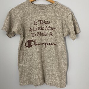 Könnte beinhalten: Ein meliertes graues Champion-T-Shirt mit dem braunen Text "It Takes A Little More To Make A Champion". Das T-Shirt hängt an einem Holzbügel vor einem schlichten Hintergrund. Es hat kurze Ärmel und einen Rundhalsausschnitt.