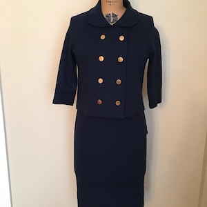 May include: A navy blue knit suit with a double-breasted jacket and a skirt. The jacket has gold buttons and a peter pan collar.