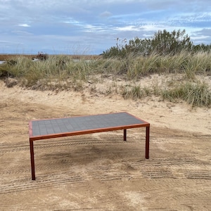 May include: A dark brown wooden coffee table with a black tiled top. The table is sitting on a sandy beach with grass and a body of water in the background.