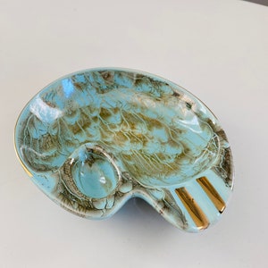 May include: Vintage ashtray with a unique design. The ashtray features a light blue and brown pattern with gold accents. It has a round indentation and two gold-lined rests. A decorative piece for a home.