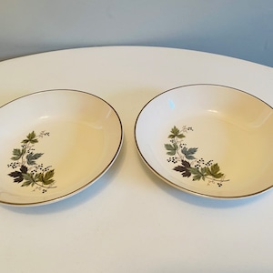 Op de afbeelding: Twee crèmekleurige ovale serveerschalen met een gouden rand. Elke kom heeft een botanisch ontwerp van groene en donkerbruine bladeren met kleine zwarte bessen. De kommen staan op een wit oppervlak.