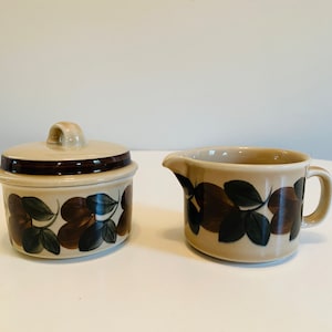 Puede incluir: Un azucarero de cerámica con tapa y una jarra para crema. Ambas piezas son beige con un estampado floral marrón y verde oscuro. El azucarero tiene forma redondeada y una pequeña asa en la tapa.