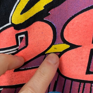May include: A black t-shirt with a colourful graphic design featuring a cartoon character with a halo. The character is wearing a yellow and black hat and has a pink and yellow body. The design is outlined in black and has a purple and blue background.