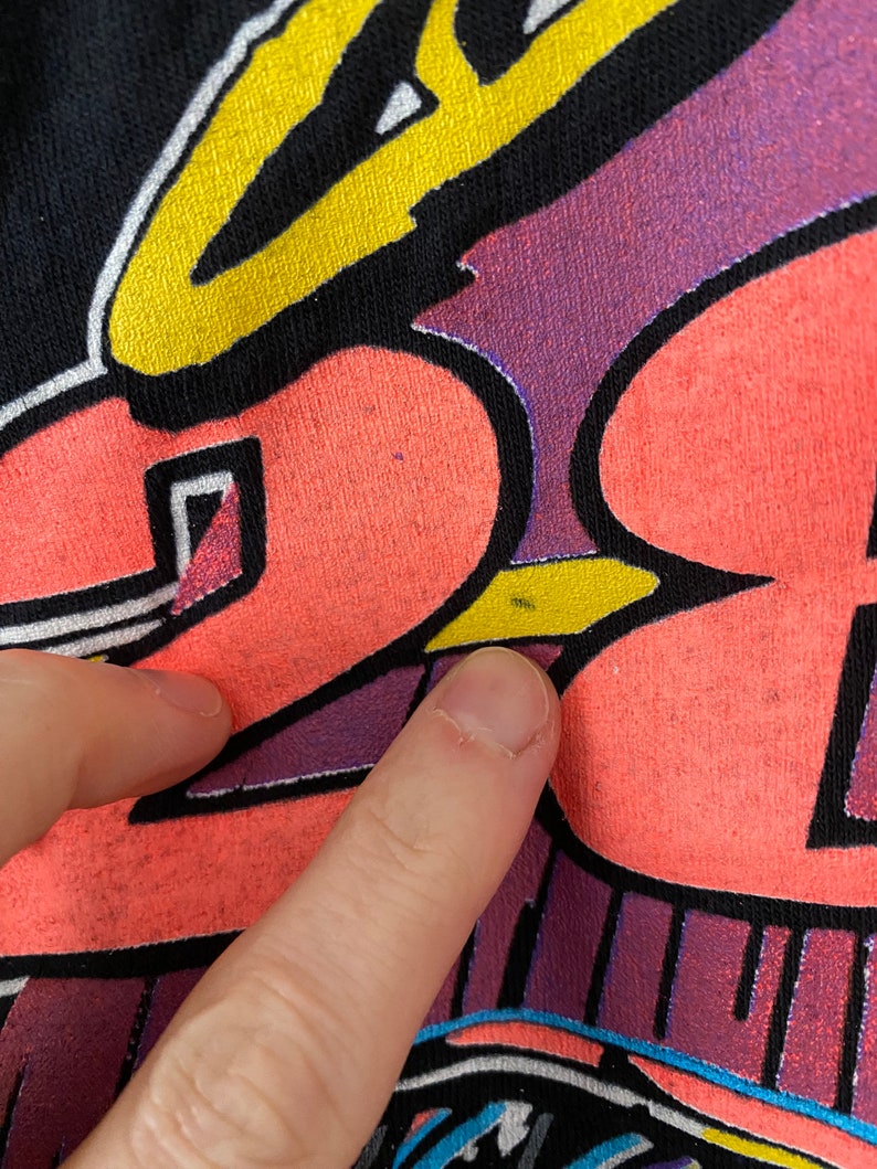 May include: A black t-shirt with a colourful graphic design featuring a cartoon character with a halo. The character is wearing a yellow and black hat and has a pink and yellow body. The design is outlined in black and has a purple and blue background.