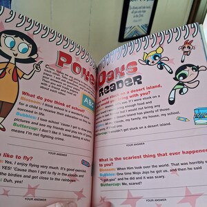May include: Open book with Powerpuff Girls illustrations and text. The book is titled "Poke Oaks Reader" and features colorful drawings of Blossom, Bubbles, and Buttercup. The pages contain questions and answers, with a pink background and star accents.