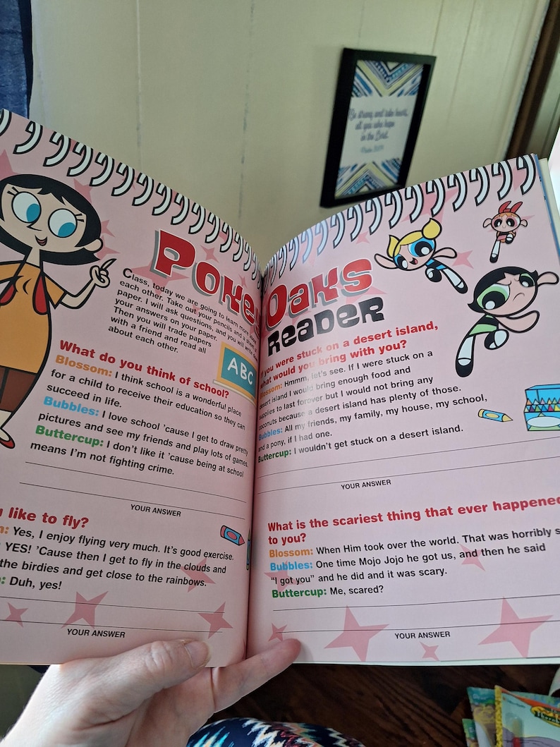 May include: Open book with Powerpuff Girls illustrations and text. The book is titled "Poke Oaks Reader" and features colorful drawings of Blossom, Bubbles, and Buttercup. The pages contain questions and answers, with a pink background and star accents.