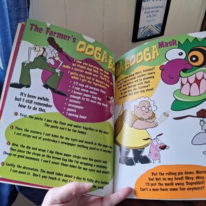 May include: Open book with colorful illustrations from the cartoon "Courage the Cowardly Dog." The pages show characters like Eustace, Muriel, and Courage, with text describing how to make a mask. The book is open, revealing the artwork.