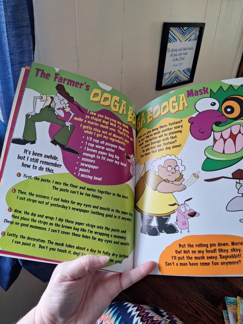 May include: Open book with colorful illustrations from the cartoon "Courage the Cowardly Dog." The pages show characters like Eustace, Muriel, and Courage, with text describing how to make a mask. The book is open, revealing the artwork.