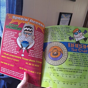 May include: Open book featuring Dexter's Laboratory characters. The left page has text and an illustration of Dexter. The right page has a code-breaking puzzle with Action Hank. The book is held open by a hand.