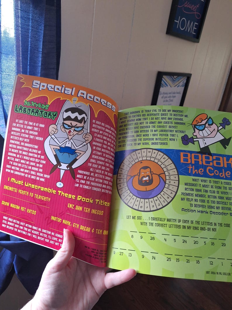 May include: Open book featuring Dexter's Laboratory characters. The left page has text and an illustration of Dexter. The right page has a code-breaking puzzle with Action Hank. The book is held open by a hand.