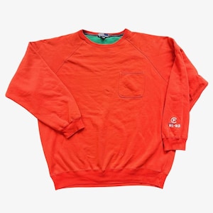 May include: A bright orange sweatshirt with a green collar and a pocket on the chest. The long-sleeved top has a small logo on the sleeve and is made of a soft material. The sweatshirt is a casual style.