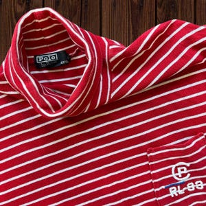 May include: Red and white striped turtleneck shirt. The shirt features a white collar and a small pocket with the embroidered text "Polo by Ralph Lauren RL-93". The shirt is made of a soft, breathable material.