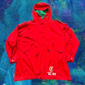 May include: A red hooded jacket with a green interior lining. The jacket features two front pockets, a drawstring at the waist, and a white logo with "RL-92" printed on the lower right side. The jacket has buttoned cuffs.
