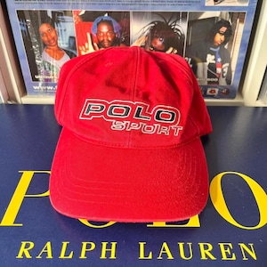 Op de afbeelding: Een rode Polo Sport baseballcap met witte en zwarte letters. De cap heeft een gebogen klep en een zes-panelen ontwerp. De hoed is gemaakt van een zacht, getextureerd materiaal. De achtergrond bevat albumhoezen.