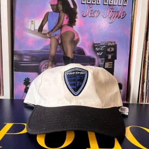 May include: A white and black baseball cap with a blue and black Polo Sport logo patch. The logo features the text "Polo Sport" and a shield with the letters "G7" and "P" inside.
