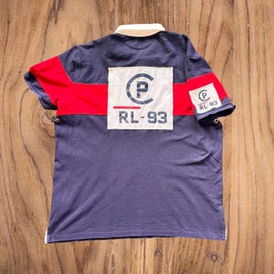 May include: Navy blue polo shirt with red accents and a white collar. The back features a large white patch with a circular logo and "RL-93" text. The sleeves have a similar logo. A classic, sporty look.