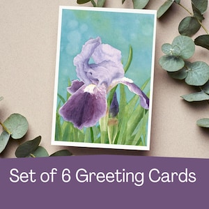 May include: A set of six greeting cards featuring a watercolor painting of a purple iris flower with green leaves against a blue background. The cards have a white border and are surrounded by eucalyptus leaves.