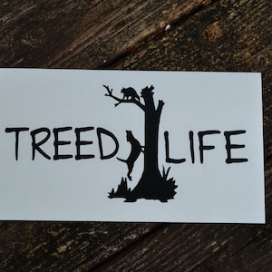 May include: A white sticker with black text and a silhouette of a dog and a cat in a tree. The text reads "TREED LIFE".
