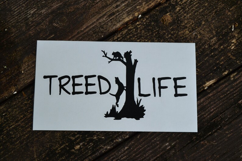 May include: A white sticker with black text and a silhouette of a dog and a cat in a tree. The text reads "TREED LIFE".