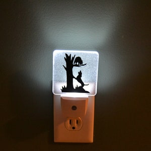 May include: A white night light with a clear plastic cover featuring a black silhouette of a cat and a dog on a tree. The night light is plugged into a standard electrical outlet.