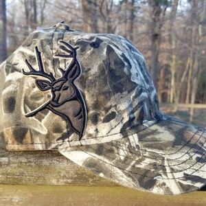 May include: Camouflage baseball cap with a black embroidered deer head logo. The cap has a curved brim and a mesh back.