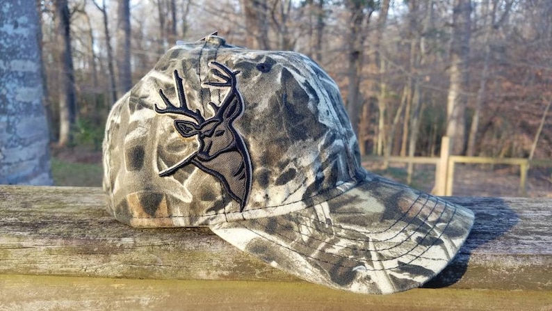 May include: Camouflage baseball cap with a black embroidered deer head logo. The cap has a curved brim and a mesh back.