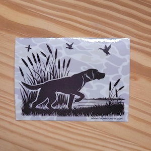 May include: A rectangular sticker featuring a black silhouette of a hunting dog in a marsh scene. The dog is in mid-stride, with cattails and ducks in the background. The website address www.coonhoundin.com is printed at the bottom.