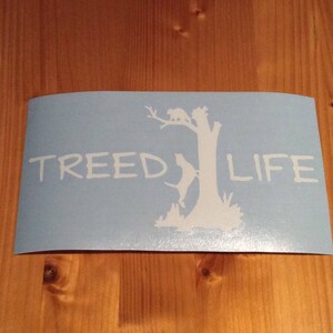 May include: A blue vinyl decal with a white silhouette of a tree with a dog and a cat climbing it. The text "Treed Life" is written in white below the tree.