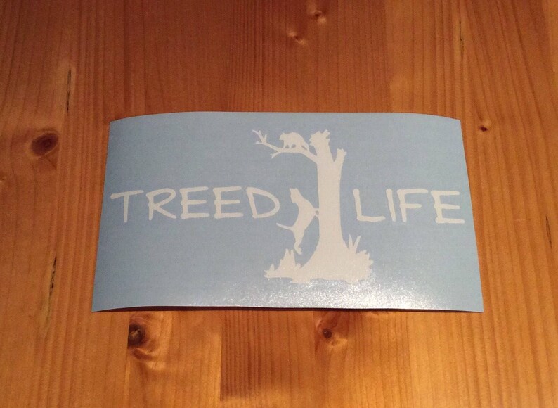 May include: A blue vinyl decal with a white silhouette of a tree with a dog and a cat climbing it. The text "Treed Life" is written in white below the tree.