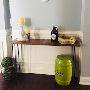 May include: A rustic console table with a dark wood top and black hairpin legs. The table is decorated with a white candle holder, a glass vase, a brown bottle with a white label, and a woven basket. A green garden stool sits below.