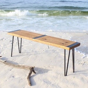 May include: A rustic wooden bench with black metal hairpin legs. The bench is made from reclaimed wood and has a natural finish. It is sitting on a sandy beach.