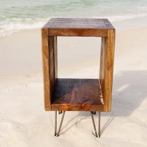 May include: A rustic wooden end table with a square open shelf and hairpin legs. The table is on a sandy beach.