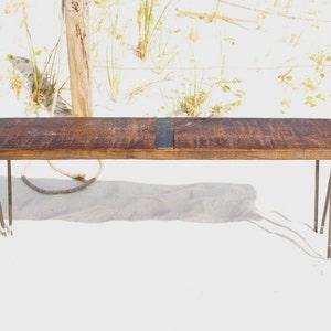 May include: A rustic wooden bench with a dark brown stain and black metal accents. The bench has hairpin legs and is sitting on a white sandy surface.