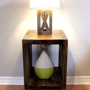 May include: A wooden end table with a white lampshade and a green and white ceramic vase. The table has a square top and three hairpin legs.