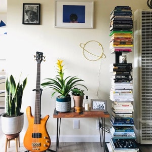 May include: A room with a bookshelf filled with books, a bass guitar, a potted plant, a small table with a potted plant, a framed picture, and a gold wire sculpture of a planet.
