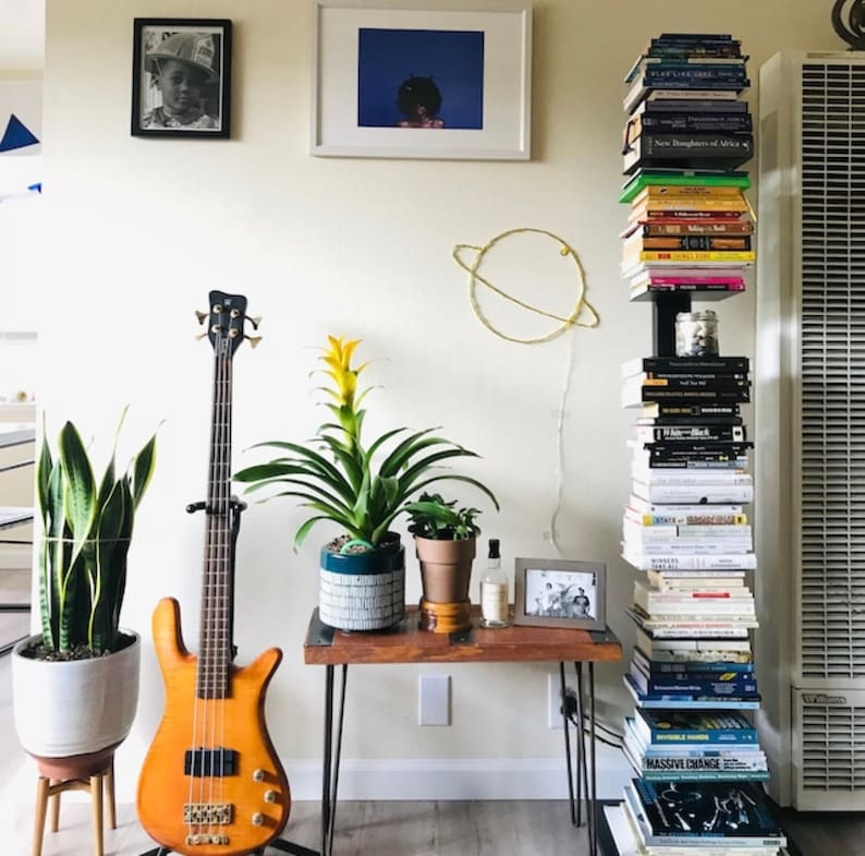 May include: A room with a bookshelf filled with books, a bass guitar, a potted plant, a small table with a potted plant, a framed picture, and a gold wire sculpture of a planet.
