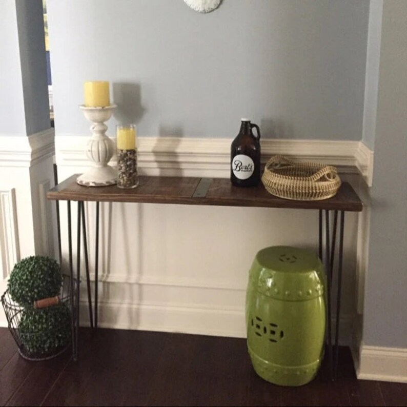 May include: A wooden console table with black hairpin legs, featuring a brown glass jar with a white label that reads "Boil's", two candles, a wicker basket, and a green ceramic stool.