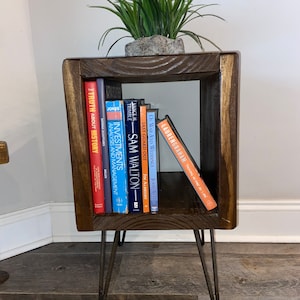 May include: A dark brown wooden end table with a square top and open shelf. The table has four hairpin legs and is decorated with a potted green plant. The shelf is filled with a variety of books, including "The Truth About History", "Investments Analysis and Management", "Last Guard Out", "Of War and Weddings", and "Lauren Graham Talking as Fast as I Can".