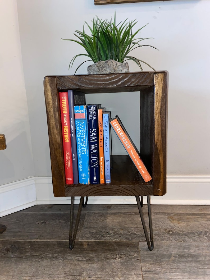 May include: A dark brown wooden end table with a square top and open shelf. The table has four hairpin legs and is decorated with a potted green plant. The shelf is filled with a variety of books, including "The Truth About History", "Investments Analysis and Management", "Last Guard Out", "Of War and Weddings", and "Lauren Graham Talking as Fast as I Can".