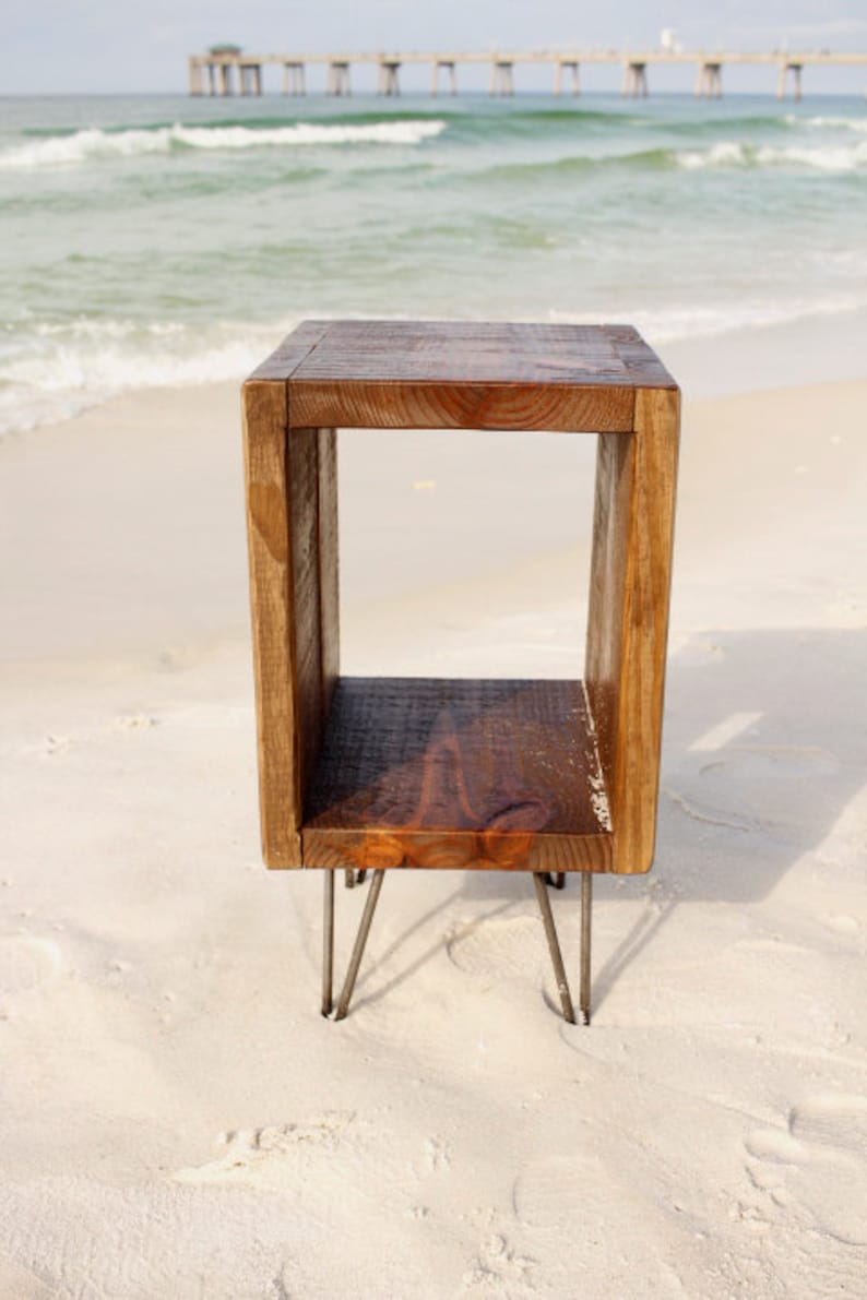 May include: A rustic wooden end table with a square open shelf and hairpin legs. The table is on a sandy beach.
