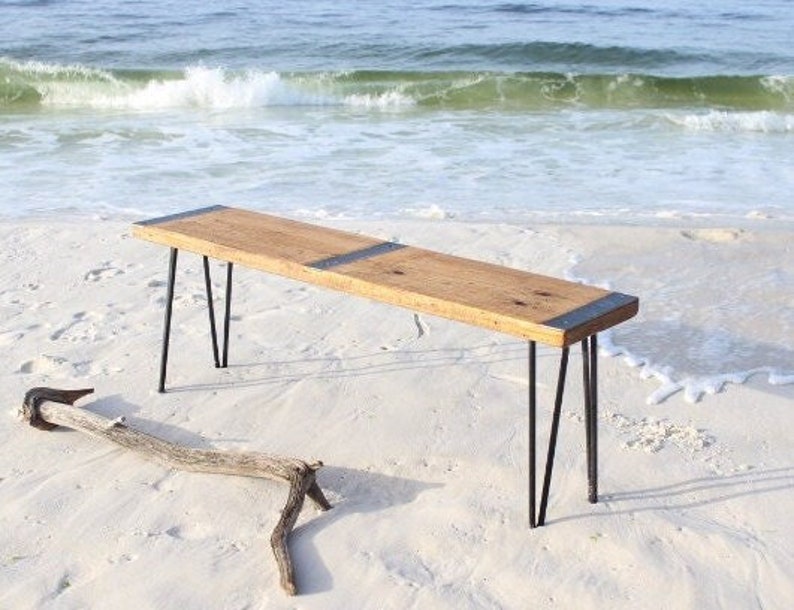 May include: A rustic wooden bench with black metal hairpin legs. The bench is made from reclaimed wood and has a natural finish. It is sitting on a sandy beach.