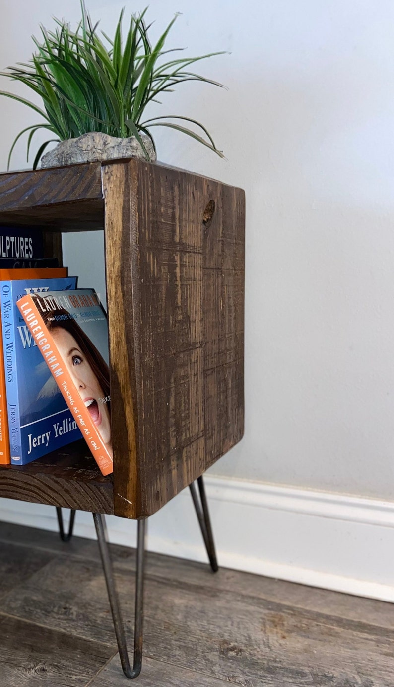 May include: A dark brown wooden end table with a shelf and hairpin legs. The table has a small potted plant on top and a stack of books on the shelf. The books include "Of War and Weddings" and "Talking as Fast as I Can" by Lauren Graham.