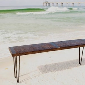 May include: A rustic wooden bench with a dark brown finish and a natural wood grain. The bench has a metal frame with hairpin legs. The bench is sitting on a sandy beach with a pier in the background.