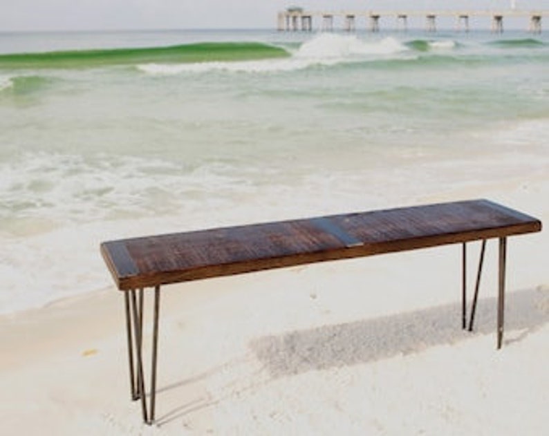 May include: A rustic wooden bench with a dark brown finish and a natural wood grain. The bench has a metal frame with hairpin legs. The bench is sitting on a sandy beach with a pier in the background.