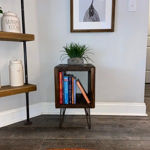May include: A dark wood end table with a single shelf, holding a stack of books. The table has hairpin legs and is sitting on a wooden floor. A potted plant sits on top of the table.