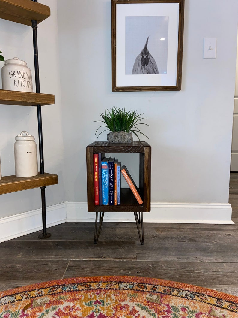 May include: A dark wood end table with a single shelf, holding a stack of books. The table has hairpin legs and is sitting on a wooden floor. A potted plant sits on top of the table.