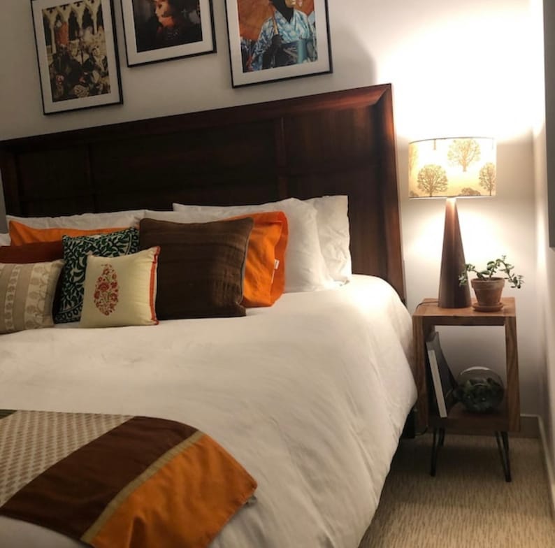 May include: A white bed with a brown headboard and orange and brown patterned bedding. A bedside table with a lamp with a brown shade featuring a tree design is next to the bed.